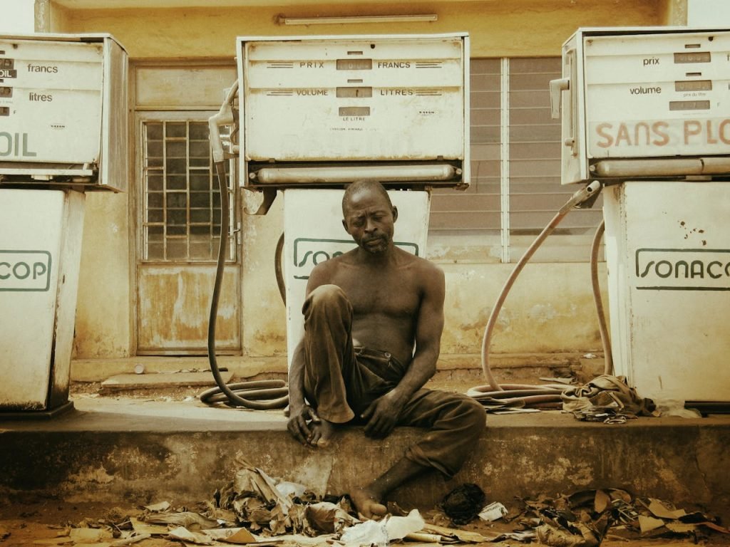 A shirtless man resting at an abandoned gas station amidst dusty surroundings.